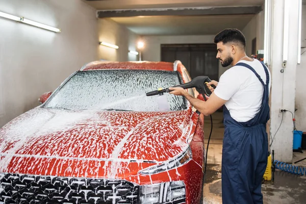 Cleaning car using high pressure water. Handsome young bearded man worker, wearing protective clothes and gloves, washing modern red car under high pressure soap water in car wash service.