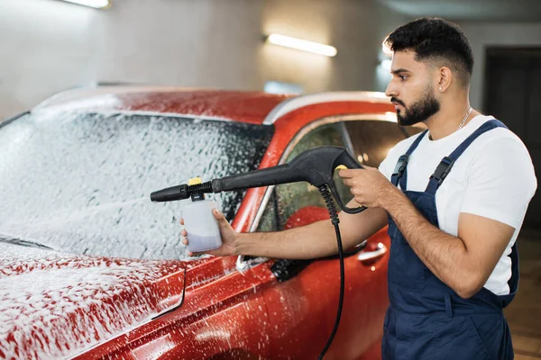Close up portrait of car wash worker, hansome young man in protective overalls, cleaning automobile with high pressure water jet at car wash, spraying the cleaning foam.