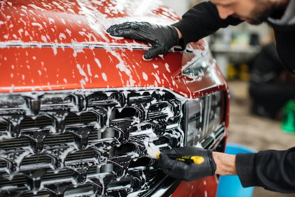 Close up photo of man hand in black protective gloves cleaning radiator grille in foam with special brush. Carwash and detailing. Washing machine at the station. Car washing concept