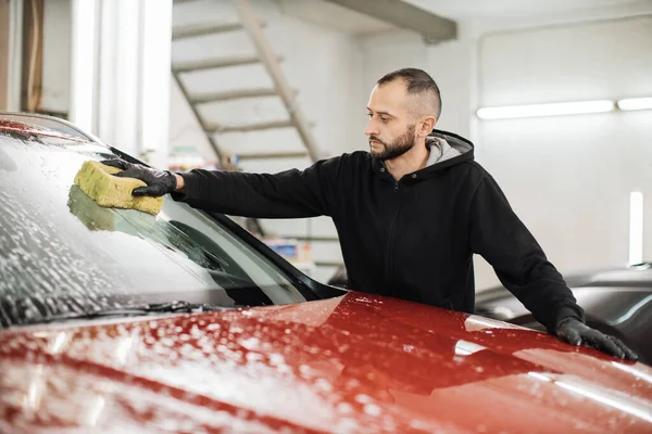 Car wash and clean with shampoo and sponge. Young handsome bearded man worker washing soapy car windshield with sponge on a car wash station.
