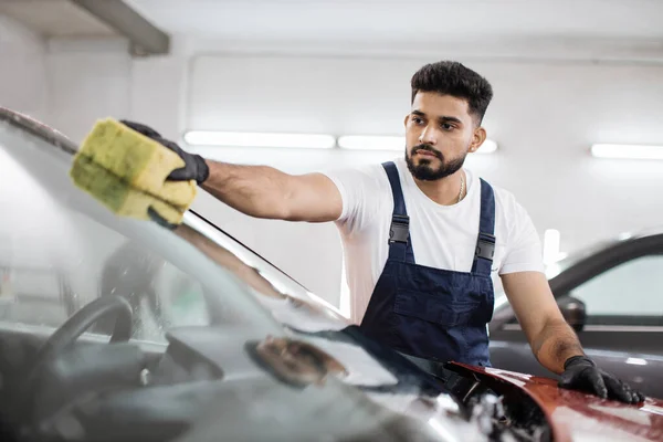 Young bearded man, car wash worker, wearing t-shirt and overalls cleaning the windshield of car with the help of special foam and sponge. Washing the car by hand, sponge and foam bubbles.