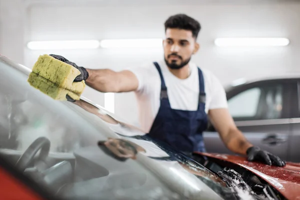Young bearded man, car wash worker, wearing t-shirt and overalls cleaning the windshield of car with the help of special foam and sponge. Selective focus on yellow sponge.
