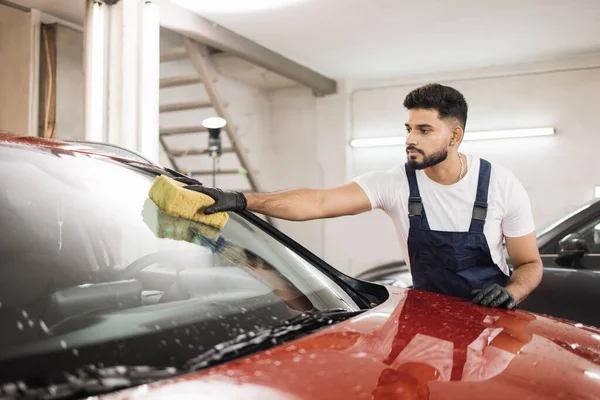 Confident young man, car wash worker, wearing t-shirt and overalls cleaning the windshield of car with the help of special foam and sponge. Washing the car by hand, sponge and foam bubbles.