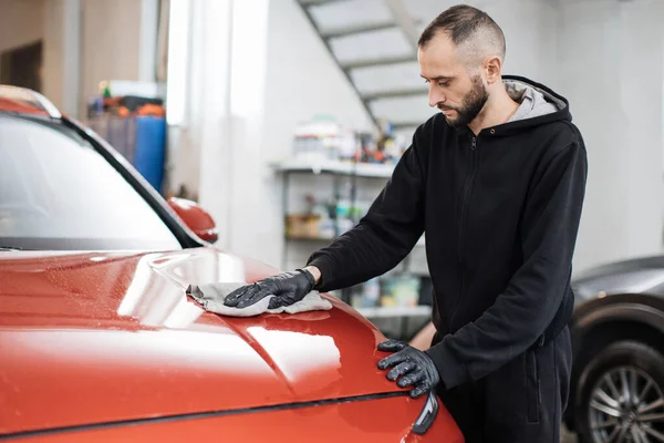 Car detailing and cleaning concept. Good-looking young male car wash worker, wearing special clothes and gloves, wipes and polishes the car hood with microfiber cloth.