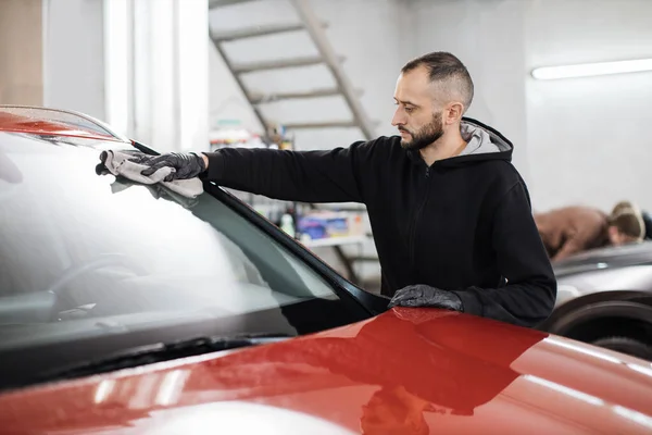 Car detailing and cleaning concept. Good-looking young male car wash worker, wearing black gloves, wipes and polishes car windshield window with microfiber cloth.