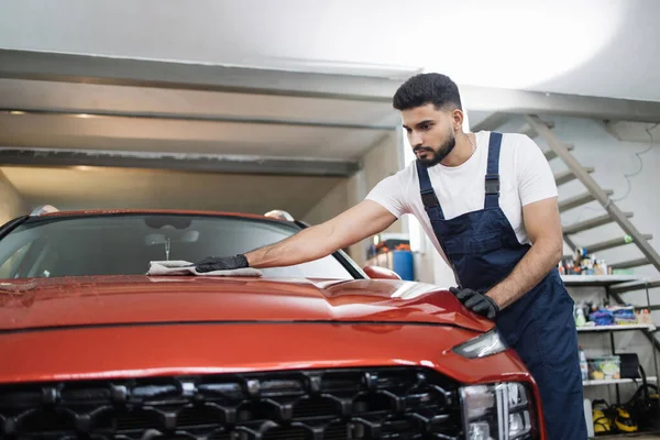 Car detailing, wash and cleaning concept. Young bearded male professional car wash worker in black rubber gloves, holding the gray microfiber and polishing the car hood of luxury car.