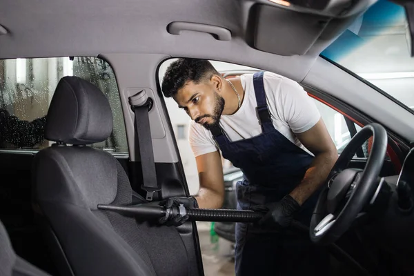 Car wash and detailing service. Bearded male worker in overalls and gloves, making chemical cleaning process with a vacuum cleaner, releasing car seat from dust and sand.
