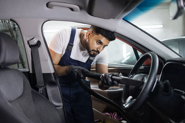 Car wash and detailing service. Bearded male worker in overalls and gloves, making chemical cleaning process with a vacuum cleaner, releasing steering wheel of the car from dust and sand.