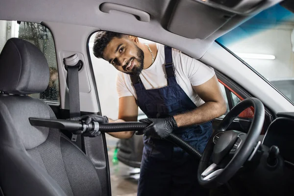 Car wash and detailing service. Bearded male worker in overalls and gloves, making chemical cleaning process with a vacuum cleaner, releasing car seat from dust and sand.