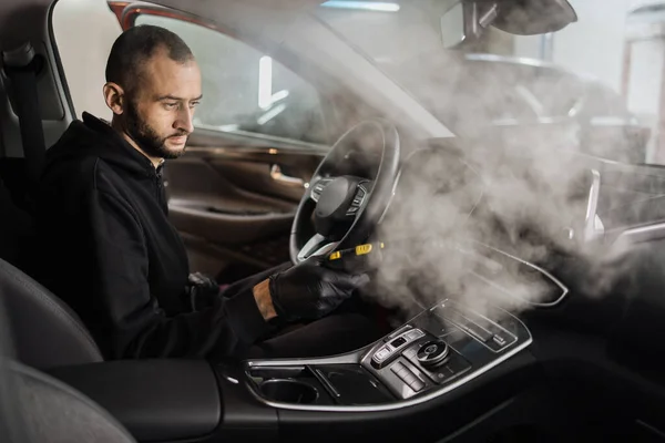 Auto cleaning service and detailing concept. Handsome bearded man in uniform cleaning interior of the car with hot steam cleaner. Selective focus on guy face.