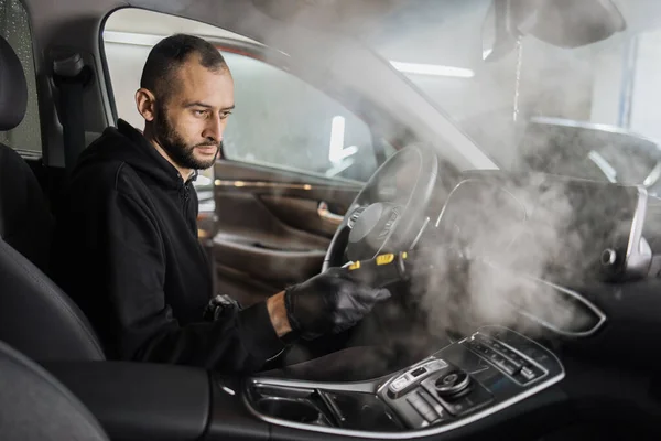 Auto cleaning service and detailing concept. Handsome bearded man in uniform cleaning interior of the car with hot steam cleaner. Selective focus on guy face.