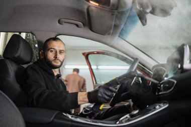 Buhar temizleyicisiyle araba salonunu temizliyorum. Profesyonel erkek kendine güvenen işçinin iç mekan temizleme, direksiyon ve kontrol paneli sıcak buhar temizleyicisi kullanıyor..