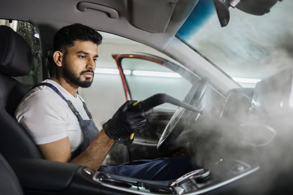 Car detailing, steam cleaning concept. Handsome man in overalls, worker of car wash center, cleaning car interior with hot steam cleaner. Car detailing concept.