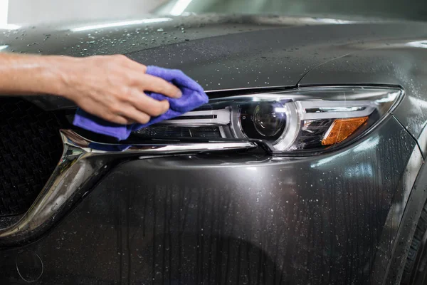 Car wash and cleaning at professional auto service station. Crooped view of hand of male worker cleaning car headlights with blue microfiber cloth.