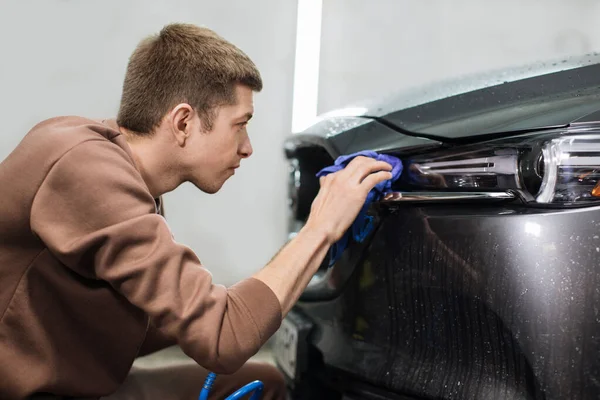 Car wash and cleaning at professional auto service station. Close up view of caucasian young male worker cleaning car headlights with blue microfiber cloth using high pressure water.