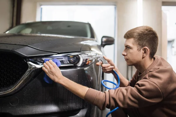 Car wash and cleaning at professional auto service station. Close up view of caucasian young male worker cleaning car headlights with blue microfiber cloth using high pressure water.