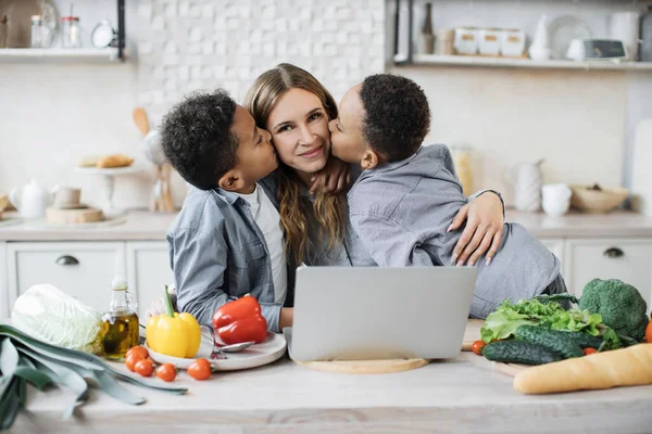 Cute preschool sons kissing their mother in light kitchen. Portrait of multinational family of african little boys and their caucasian mom reading healthy salad recipe on laptop.