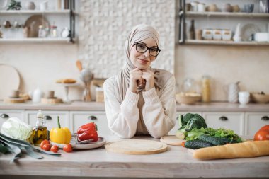 Genç tesettürlü tesettürlü Müslüman kadın taze sebzeler ve baget ekmeğiyle modern hafif mutfakta oturmuş taze vejetaryen malzemelerinden diyet salatası yapmaya hazırlanıyor..
