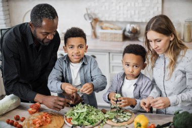 Gülümseyen çocukların anne ve babalarına yemek ya da öğle yemeği için yardım etmesi, marul yapraklarını kâseye dökmesi. Çok uluslu aileler ve oğulları mutfakta yemek pişirip birlikte salata hazırlıyorlar..