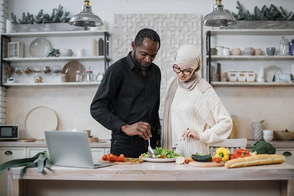 Attractive african man and young muslim woman holding spoons and mixing ingredients while preparing delicious healthy salad for breakfast or dinner in the light kitchen at home.