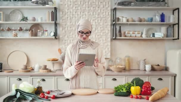 自宅で夕食のためのヒジャーブ料理サラダで陽気な若いイスラム教徒の女性は デジタルタブレットを見て 食品ブログを読んで キッチンのインテリア コピースペース 料理のコンセプト — ストック動画
