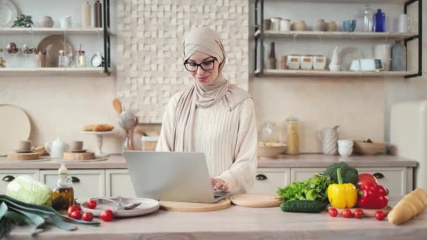 健康饮食 科技与饮食理念 一位迷人的穆斯林妇女站在桌旁准备早餐蔬菜沙拉 在家里厨房用笔记本电脑观看 — 图库视频影像