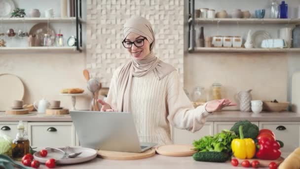 現代のキッチンでおいしいと健康的な食べ物を調理するイスラム教徒の女性は ラップトップを見て オンラインクラス レッスンやソーシャルネットワークのフォロワーとのビデオ通話を持っている間 自宅でインターネットのメディア — ストック動画
