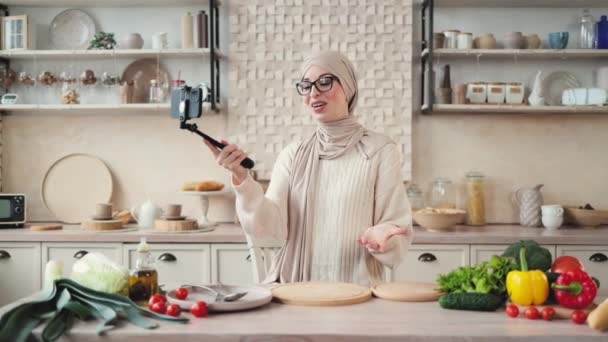 ムスリム女性がセルフィースティックを使用してビデオ通話を持ち オンラインクラスの健康的なビーガン料理を提供しています幸せな若い女性ブロガーは 彼女のフォロワーに新鮮な野菜と夕食を準備する方法を示す — ストック動画