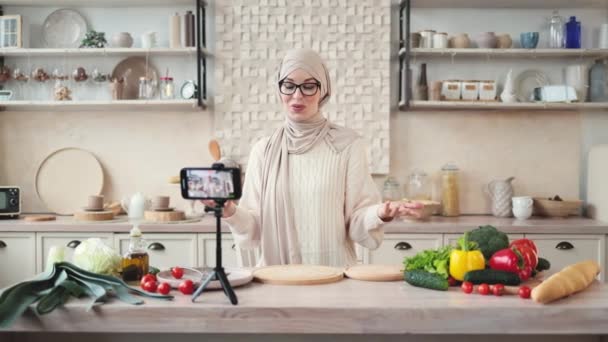 スマートフォンで食事や食事のブログのための新しいビデオを記録する興奮したイスラム教徒の女性 キッチンに立って 携帯電話のカメラでサラダの独自のレシピについて話す 選択的な焦点 — ストック動画