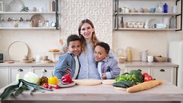 健康的食物 家庭休息的概念 年轻漂亮的母亲和两个孩子在厨房做饭的时候 一边用新鲜蔬菜做沙拉 一边抱着孩子 一边笑着看着摄像机 — 图库视频影像