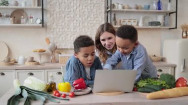 Çok uluslu Afrikalı küçük oğulların portresi ve laptopta sağlıklı salata tarifi okuyan beyaz anneleri mutfaktaki ahşap masada oturuyor, eğleniyor, hafta sonunun tadını çıkarıyor..