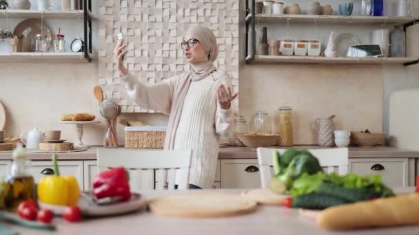 夕食の準備をしているかなり若いアラビア人の女性野菜レシピを検索食事メニュー料理アプリスマートフォンを使用してスマートフォン笑顔イスラム教徒の女性は 自己撮影を行うか 健康的なビーガン料理を調理中にビデオ通話を持っています — ストック動画