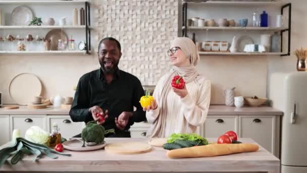 若い魅力的なカップルは新鮮な野菜からサラダを準備するのが大好きです ハンサムなアフリカ人男性ジャグリングトマトとイスラム教徒の魅力的な女性が一緒に夕食を調理し 軽いキッチンで楽しんで — ストック動画