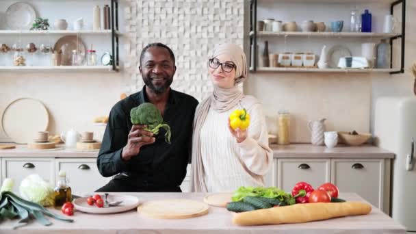 魅力的なアフリカ系アメリカ人男性とイスラム教徒の若い女性が一緒に夕食を調理しながら新鮮な野菜ブロッコリーとコショウを保持し 新しい軽いキッチンで楽しみを持っています — ストック動画