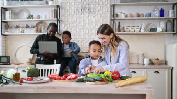 キッチンでおいしい料理を作るかなりの母親と息子 幸せな父親と子供の男の子がラップトップコンピュータを使用して時間を過ごしている間に調理するために素敵な子供を教えています 健康的な食事と夕食の準備 — ストック動画