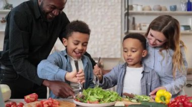 Genç ebeveynler Afrikalı küçük şirin oğullara dilimlenmiş sebzelerle sağlıklı vejetaryen yemeği yapmayı öğretiyor. Annem ve babam salata tarifini paylaşıyor, çok ırklı aile sohbetleri yapıyor mutfakta eğleniyorlar..