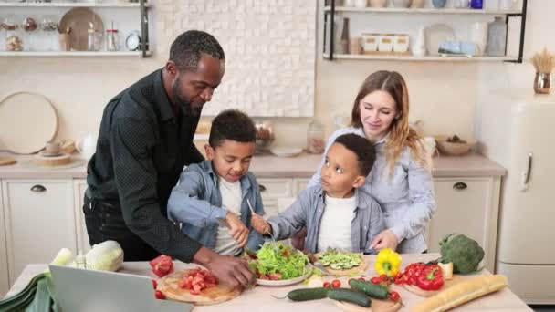 年轻的父母教非洲小可爱的儿子们用切片蔬菜准备健康的素食 妈妈和爸爸分享沙拉配方 多种族家庭在厨房聊天享受时光 — 图库视频影像