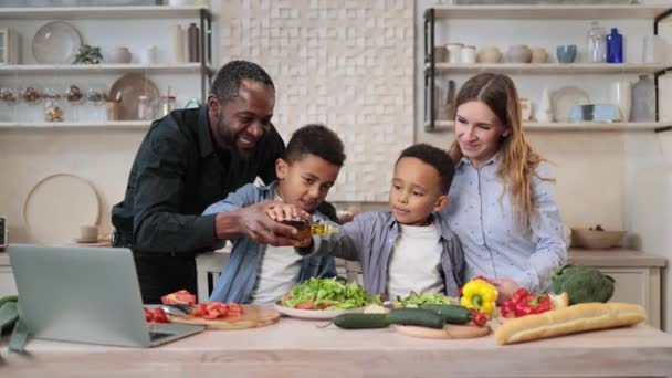 爸爸和小儿子们在厨房里一起做饭的时候 在沙拉里加橄榄油的形象 快乐的多种族父母和小男孩在家里准备健康食品很开心 — 图库视频影像