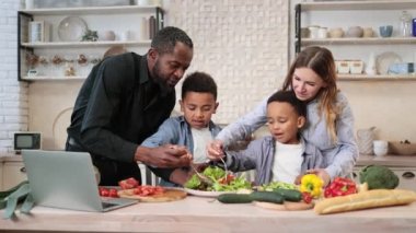 Genç mutlu anne, baba ve küçük oğullar mutfakta birlikte yemek pişirirken salataya zeytin yağı ekliyorlar. Mutlu çok ırklı ebeveynler ve küçük çocuklar evde sağlıklı yemekler hazırlarken eğlenin..