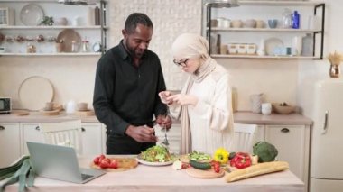 Afro-Amerikalı adam kaşıkla çatal tutuyor, kasede malzemeleri karıştırıyor, lezzetli sağlıklı salata hazırlıyor, mutfakta masada yemek yiyor, karısının yanında sosyal medya için fotoğraf çekiyor..