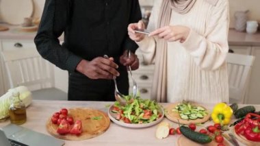 Kaşık ve çatal tutan Afrikalı adamın elleri, kasede malzemeleri karıştırma, lezzetli sağlıklı salata hazırlama, mutfakta masada yemek hazırlama, karısının sosyal medya için fotoğraf çekmesi,