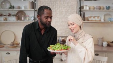 Neşeli evli çift, mutfakta sağlıklı yemek pişirirken dizüstü bilgisayar kullanıyor, tesettürlü müslüman genç kadın kocasını taze sebzelerle çatalla besliyor..