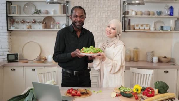 魅力的なアフリカ系アメリカ人男性とイスラム教徒の若い女性が一緒に夕食を調理しながら 新鮮な野菜と健康的なサラダとボウルを保持し 新しい軽いキッチンで楽しみを持っています — ストック動画