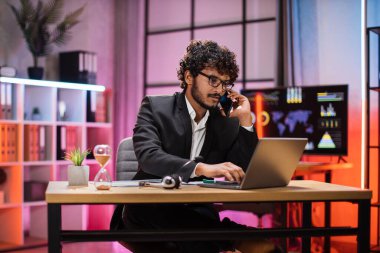 Curly bearded businessman having mobile conversation while sitting at table and working on wireless laptop. Working process at office of young guy at evening time.