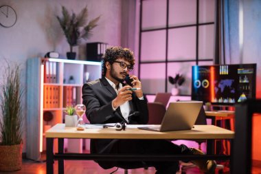 Serious confident curly bearded businessman, office manager, wearing suit using laptop while having phone call, making financial report, writing on paper, working at night.