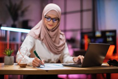 Focus in hand with pen. Attractive confident muslim business woman, office manager, wearing hijab using laptop while making financial report while writing on paper working at night.