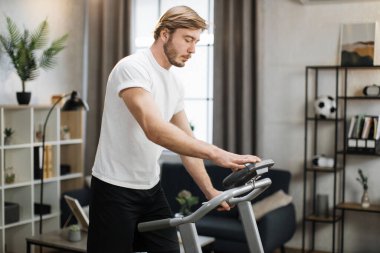 Side view of healthy athlete having cardio workout indoors. Young bearded blond man training at home while running at treadmill, tapping on control panel screen of machine.
