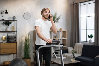 Side view of attractive sporty man in white t-shirt working out in treadmill while having phone call with his relatives or friends. Home activity, lifestyle concept.