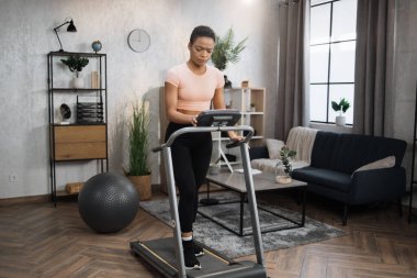 Full length portrait of young sportswoman training at home gym using treadmill. Pretty fit woman tapping on screen of machine to choose speed and training mode.
