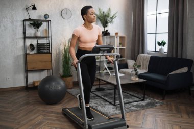 Young attractive sporty african american woman in sportswear having morning workout at modern light apartment. Healthy athletic female jogging while training at treadmill.